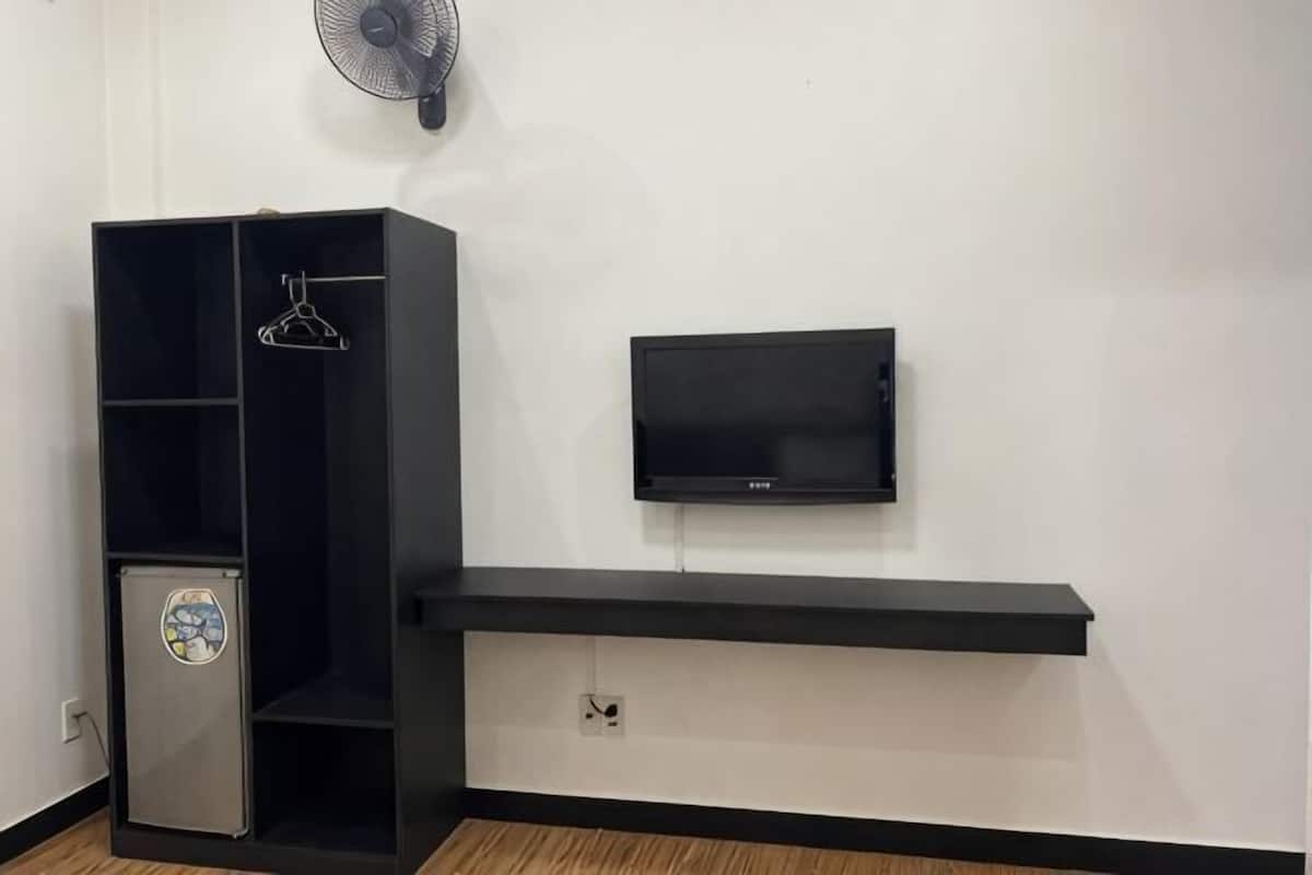 A room with a black entertainment unit, a small refrigerator, a mounted TV, and a wall-mounted fan.