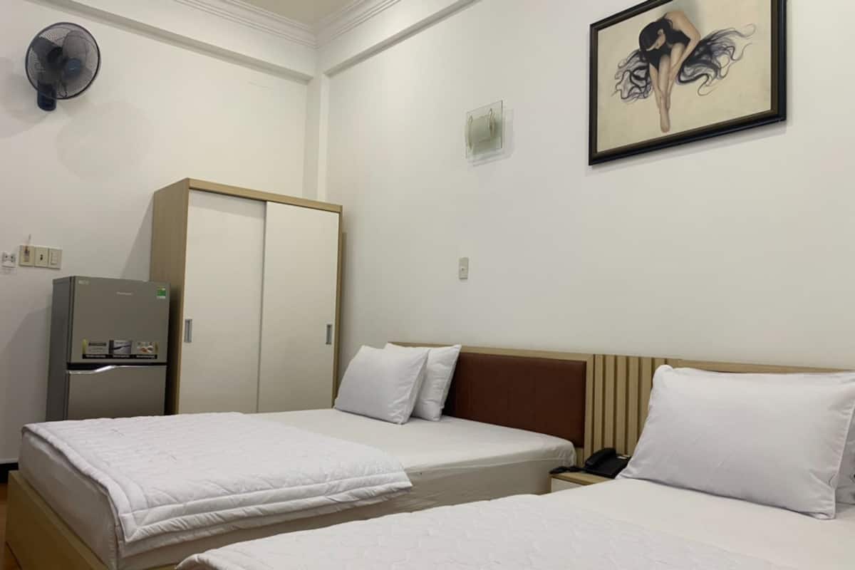 A hotel room with two beds, a wardrobe, a refrigerator, a fan, and a painting on the wall.