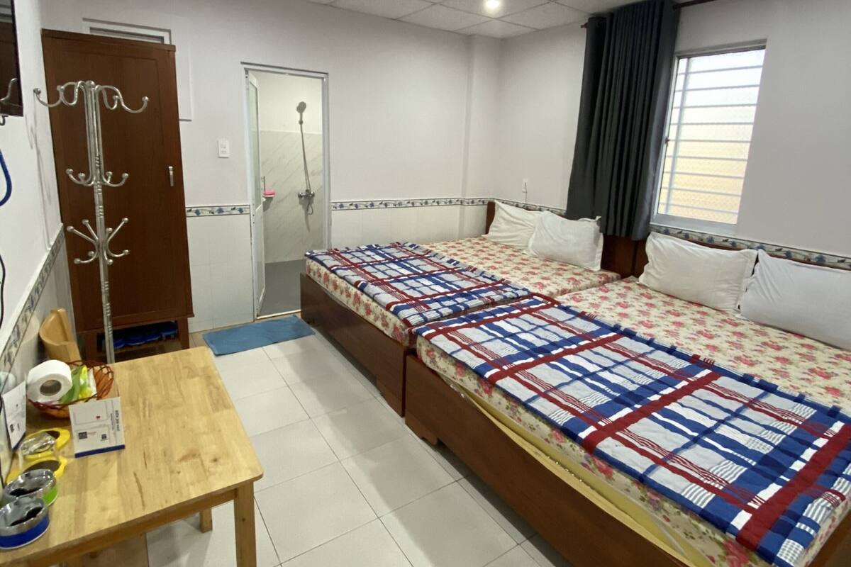 A bedroom with a large bed, a wooden table, a desk, a mirror, a ceiling fan, a window with curtains, and a door.