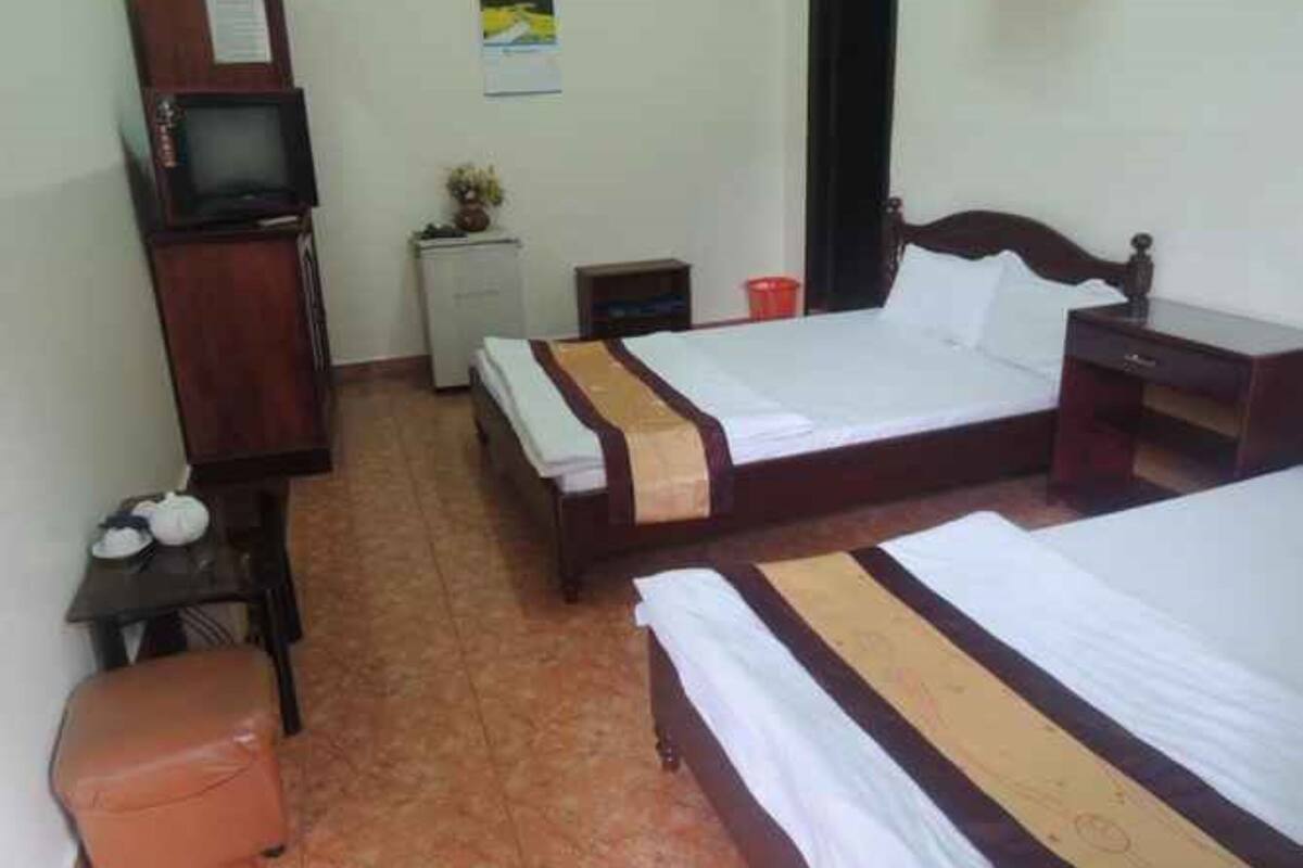 A hotel room with two single beds, a wooden wardrobe, a small desk, a chair, a nightstand, a red trash bin, and a wall-mounted TV.