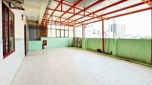 A spacious balcony with a red metal roof structure, tiled floor, and a view of a cityscape.