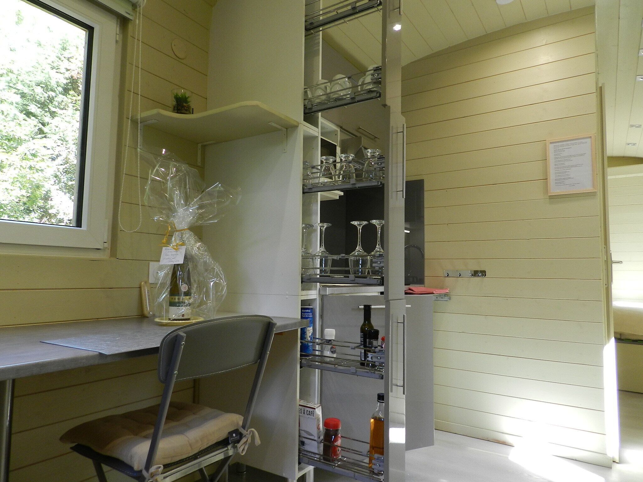 A room with a metal shelf unit containing glassware and bottles, a chair, and a window with a view of trees.