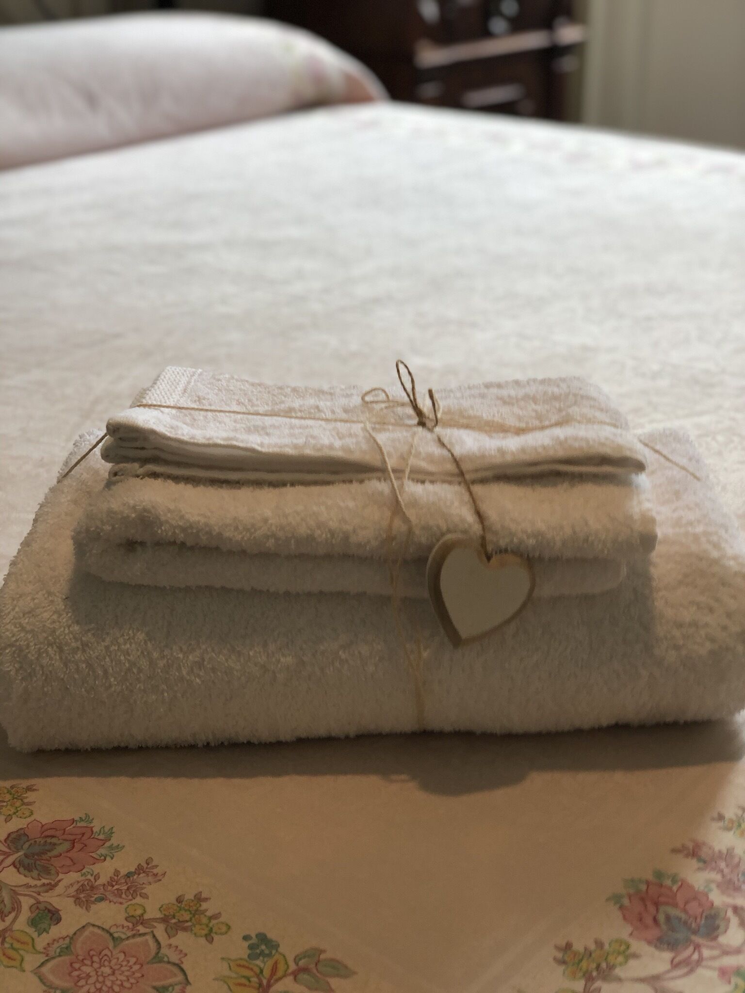 Ein ordentlich gefaltetes weißes Handtuch mit einem herzförmigen Anhänger auf einem Bett mit einem blumigen Überwurf.