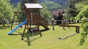 Un parque infantil de madera con un tobogán y estructuras para escalar, rodeado de césped y árboles.