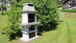 Una parrilla de piedra al aire libre con hogar y leña almacenada debajo, rodeada de follaje y un edificio al fondo.