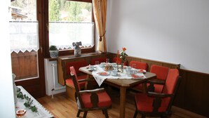 Un comedor con una mesa de madera dispuesta para cuatro personas, rodeada de sillas tapizadas en rojo. Se ve una ventana con cortina, y un radiador montado en la pared.