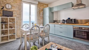 A modern kitchen with stone walls, a dining area with wicker chairs, and a view of the sea. - Charmant T2 Vue Mer Décoration Soignée (Saint-Aubin-sur-Mer)