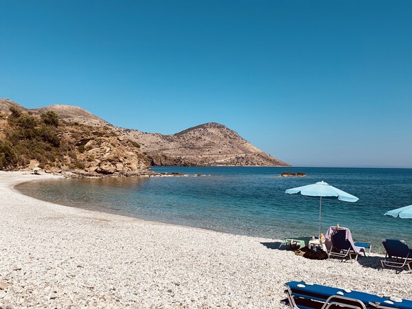 Una playa de guijarros con agua azul cristalina, algunas sombrillas y sillas.
