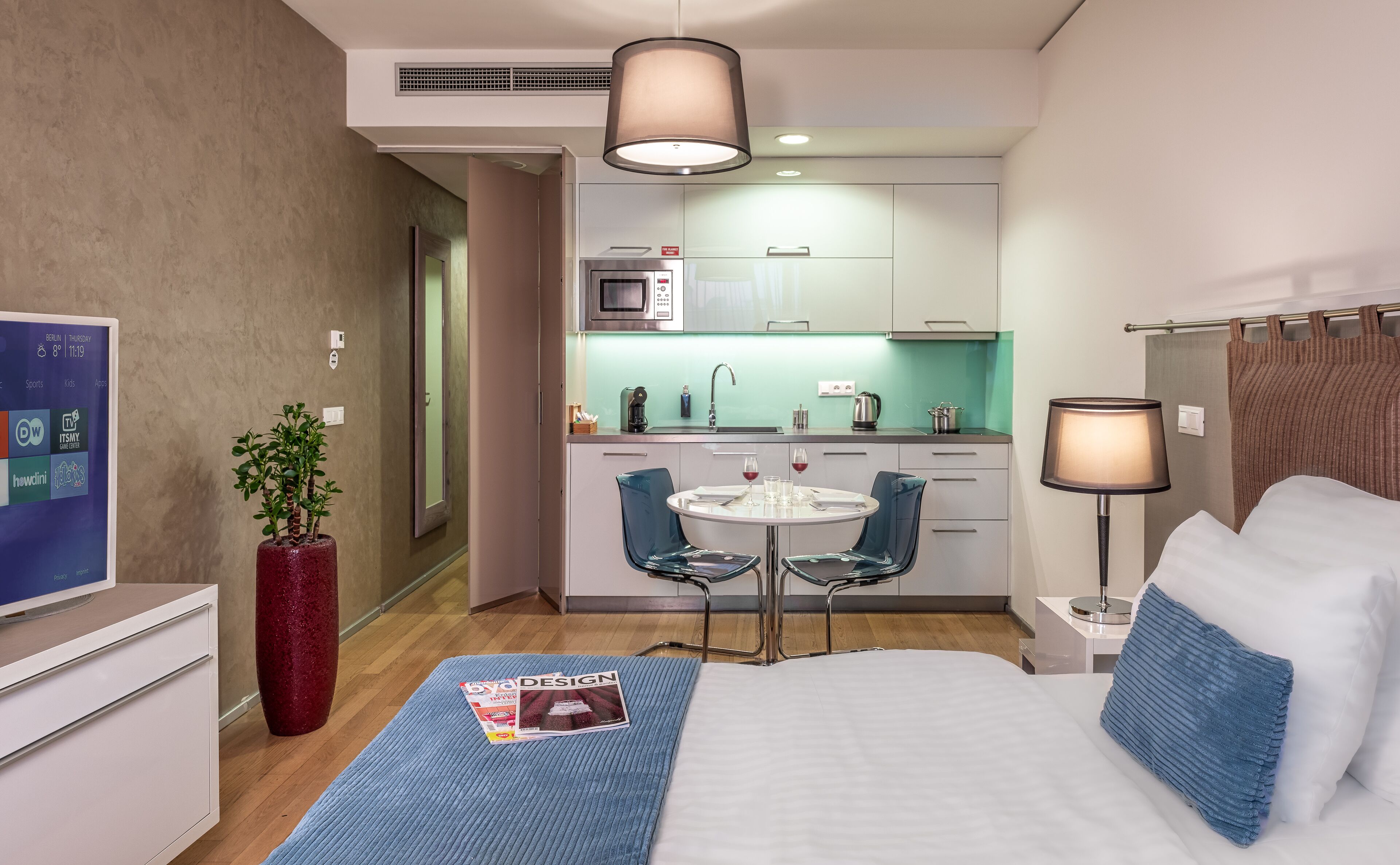A hotel room with a bed, a desk, a chair, a small kitchen area, and a TV.