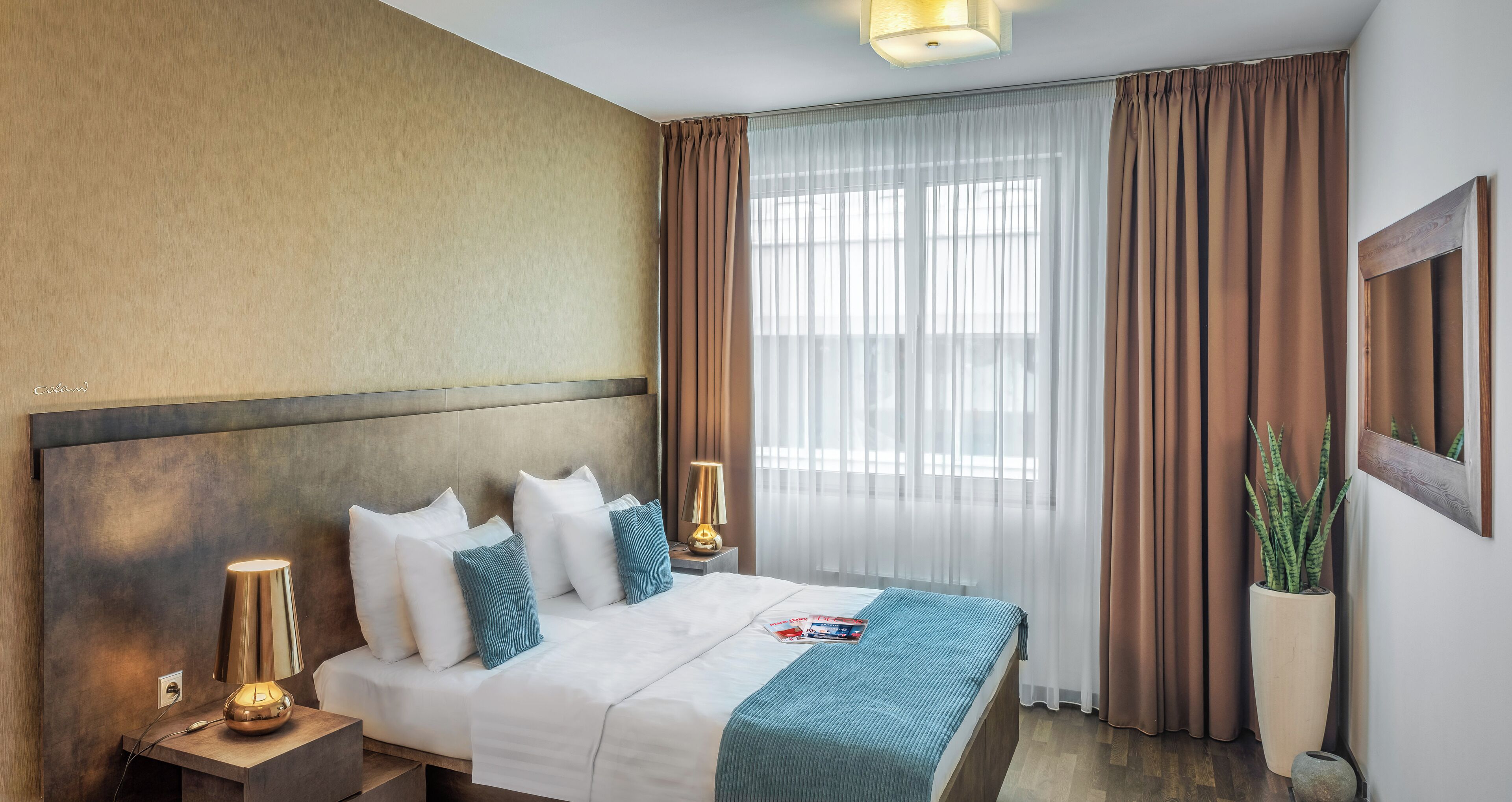 A hotel room with a large bed, a bedside lamp, a nightstand, a potted plant, and a window with curtains.
