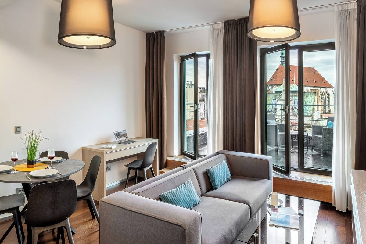 A modern hotel room with a dining area, a sofa, and a large window with a city view.