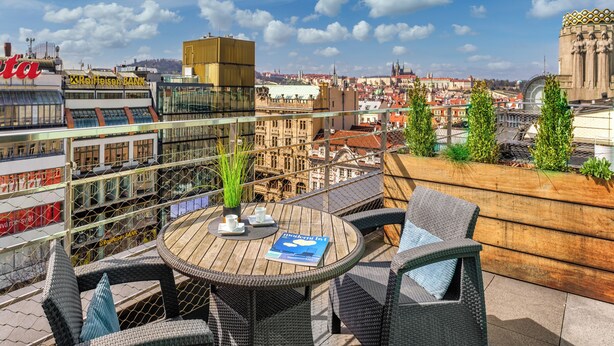 A rooftop terrace with wicker chairs, a round wooden table, and a view of a cityscape including historic buildings and a modern bank.
