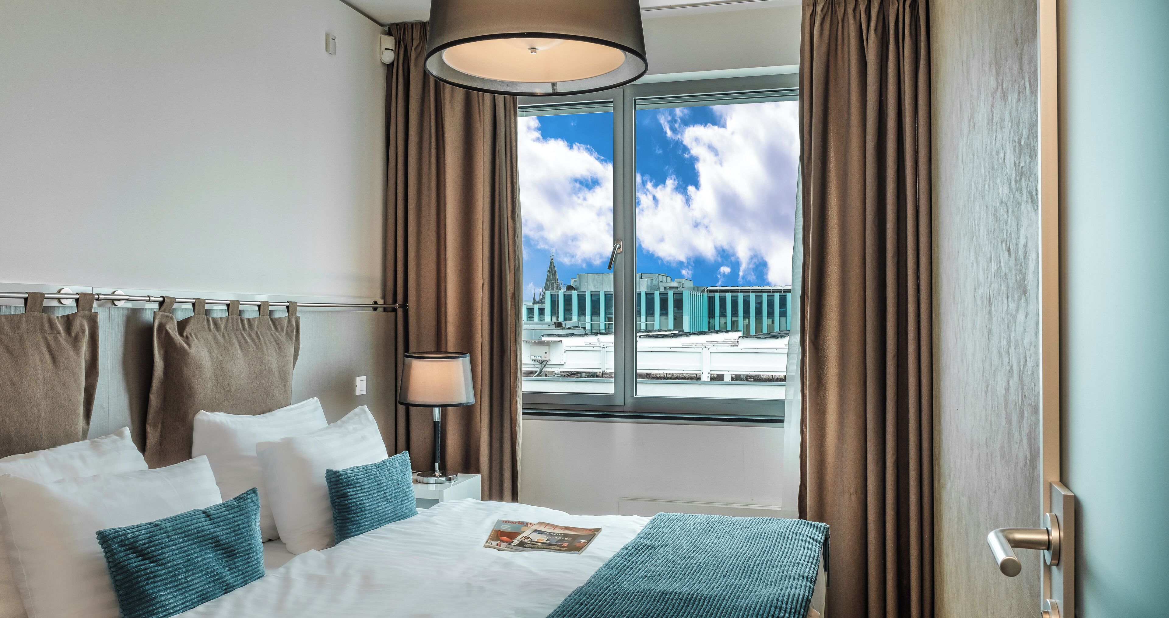 A hotel room with a large bed, a window with curtains, a lamp, and a view of a building outside.