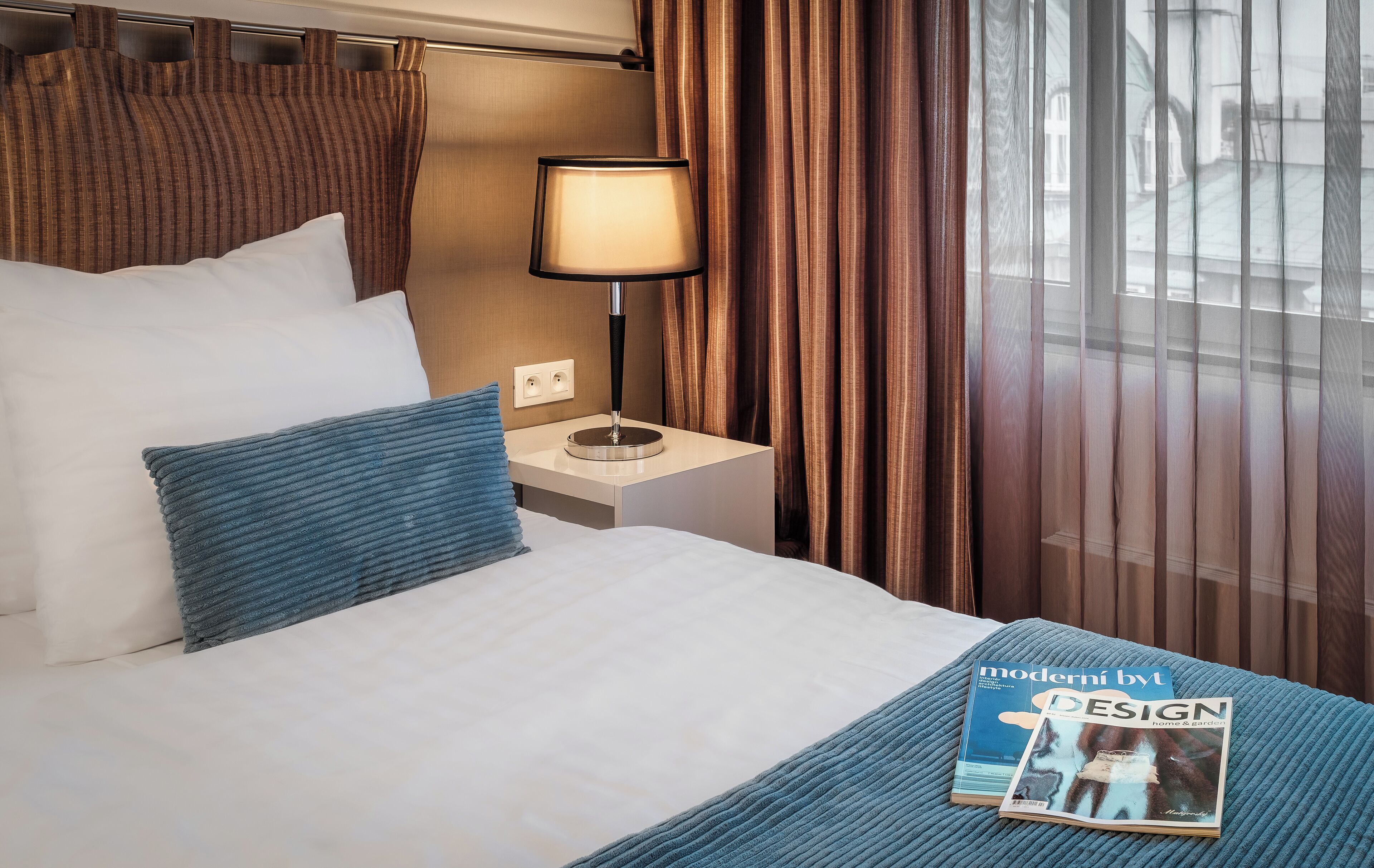 Una habitación de hotel con cama, mesita de noche con lámpara, una revista sobre la cama y ventana con cortinas.