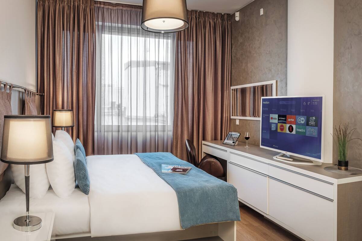 A hotel room with a bed, bedside lamps, a desk with a flat-screen TV, and a large window with curtains.