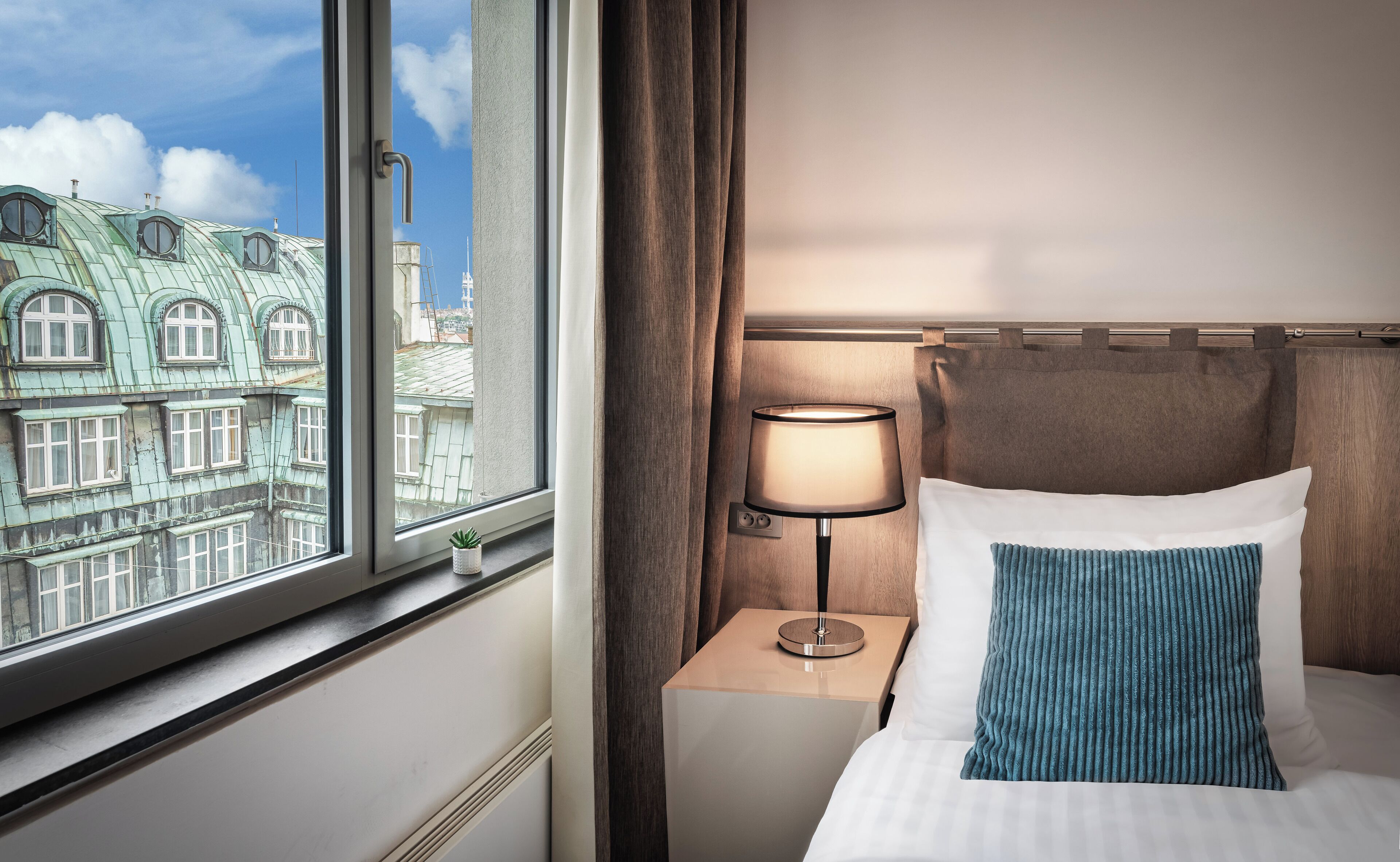 Ein Hotelzimmer mit einem Bett, einem Nachttisch mit Lampe, einem Fenster mit Blick auf historische Gebäude und einer kleinen Pflanze auf dem Fensterbrett.