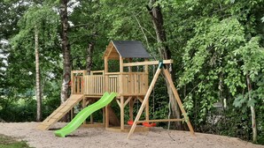 Eine Holzspielkonstruktion mit Rutsche und Schaukeln, umgeben von Bäumen und Sand.