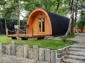 Een houten accommodatie in de vorm van een pod, met een zwart dak, een klein terras met een tafel en stoelen, en een stenen muur op de voorgrond.