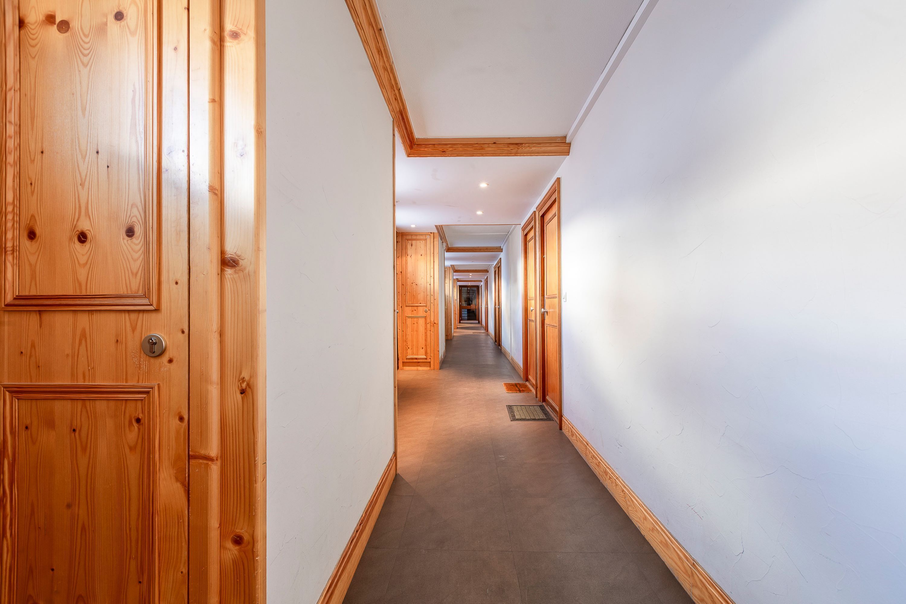 Un pasillo largo y estrecho con puertas y paredes de madera, que conduce a una zona central con una puerta al final.