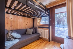 Una habitación acogedora con suelo de madera, un sofá gris con cojines estampados, una ventana con vistas a un paisaje nevado y un radiador.