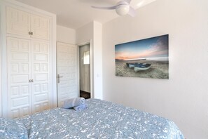 Quarto com cama, ventilador de teto, pintura de uma cena de praia e portas duplas brancas.