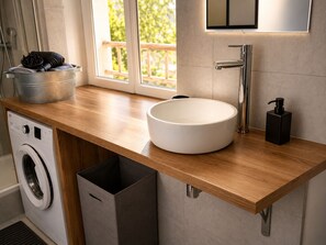 A modern bathroom with a wooden countertop, a white sink, a faucet, a washing machine, and a trash bin.