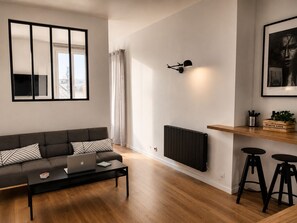 A modern living room with a sofa, a coffee table with a laptop, a radiator, a wall-mounted light, and a shelf with a plant and utensils.