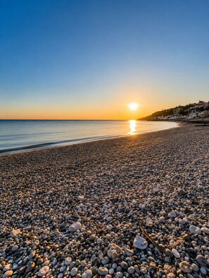 A pebbled beach with a sunset, calm sea, and buildings in the background.