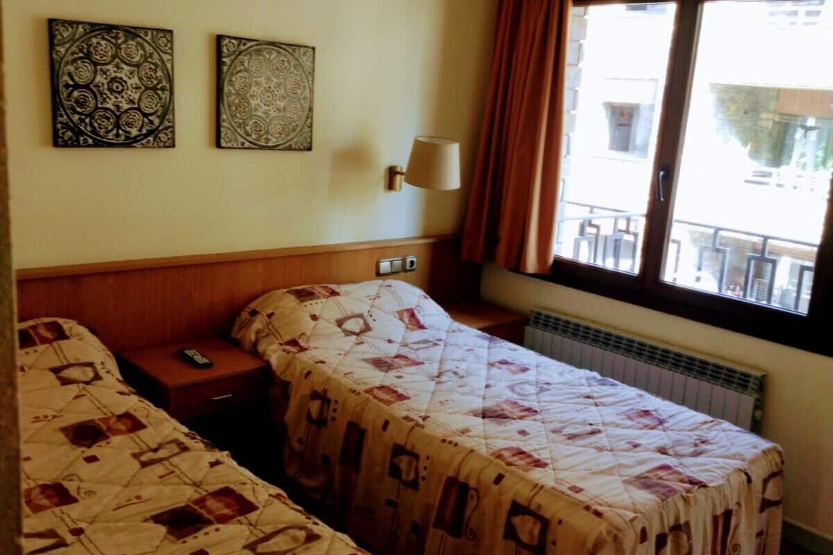 Habitación de hotel con dos camas individuales, una mesita de noche, una lámpara y una ventana con cortinas.