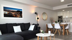 A modern living room with a black sofa, a dining area with white chairs, a kitchen in the background, and a framed cityscape picture on the wall.
