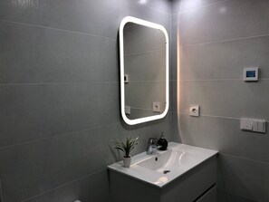 A modern bathroom with a rectangular mirror, a white sink, a potted plant, and a light switch on the wall.