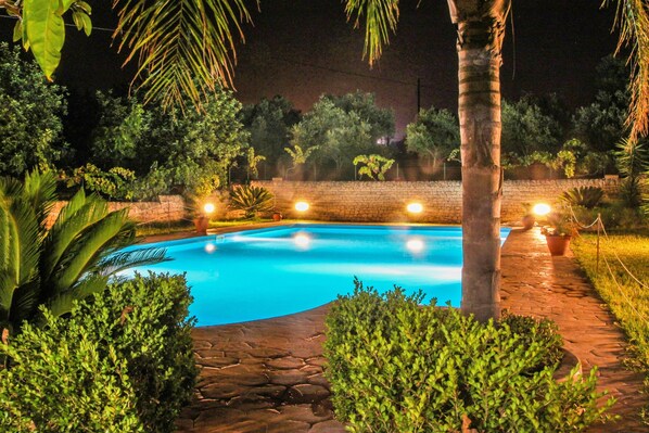 A nighttime swimming pool surrounded by greenery and stone pathways.