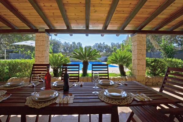 An outdoor dining area with a view of a pool, surrounded by greenery. - Villa Torres (Scicli)