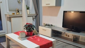 Un salon moderne avec un canapé blanc, un plaid rouge, une table basse, un téléviseur et une petite étagère ornée d’objets décoratifs.