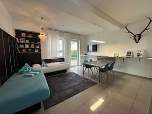 A modern living room with a sofa, a dining table with chairs, a kitchen area, and a mounted deer skull on the wall.