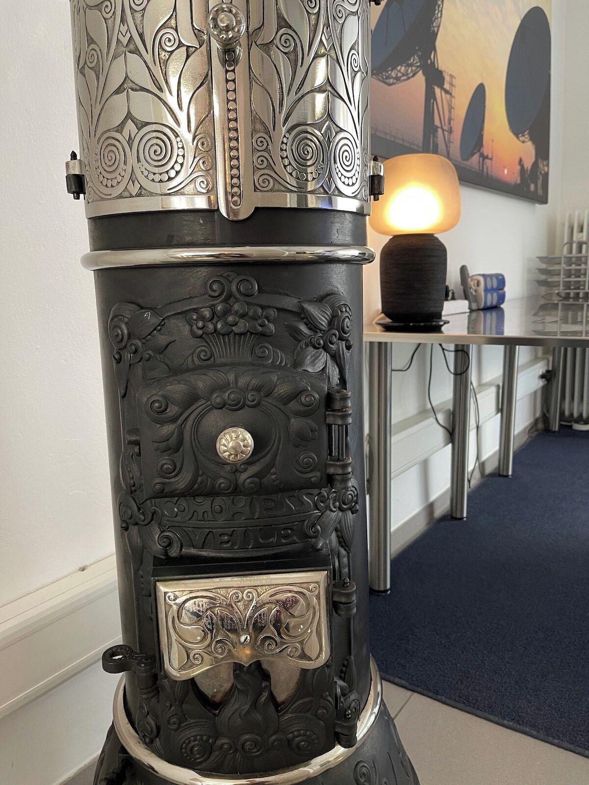 A vintage stove with intricate metalwork and a brass front plate, set in a modern kitchen with a dining table and a lamp in the background.