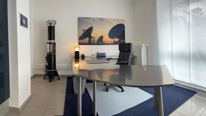 Una oficina moderna con una mesa de vidrio, una silla negra, una lámpara de escritorio y un cuadro con antenas satelitales.
