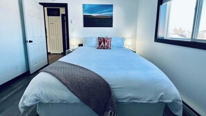 A modern bedroom with a large bed, a view of the outdoors through a window, and a door leading to another room.