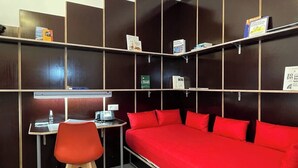 Een kamer met een rode bank, een bureau met stoel, en planken met boeken en een thermostaat.