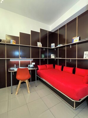 A room with a red sofa, a desk with a chair, and shelves with books and a thermostat.