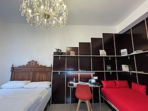 A hotel room with a wooden headboard bed, a desk with a chair, a red sofa, a chandelier, and built-in shelving with books and a phone.