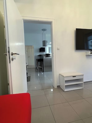 A modern living room with a television, a bench, and a door leading to a kitchen area.