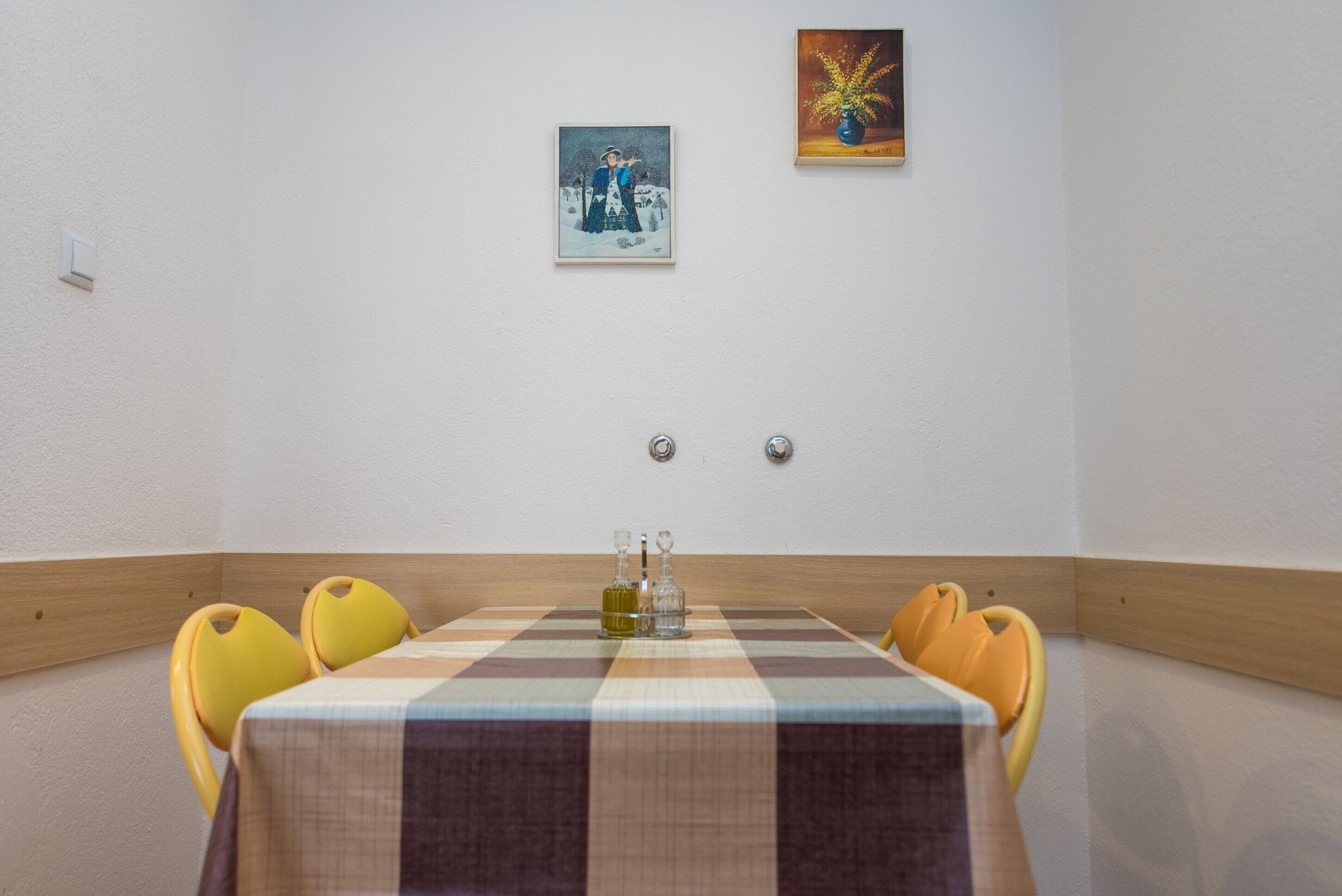 A dining area with a checkered tablecloth, four yellow chairs, two wall-mounted light switches, and two framed artworks.