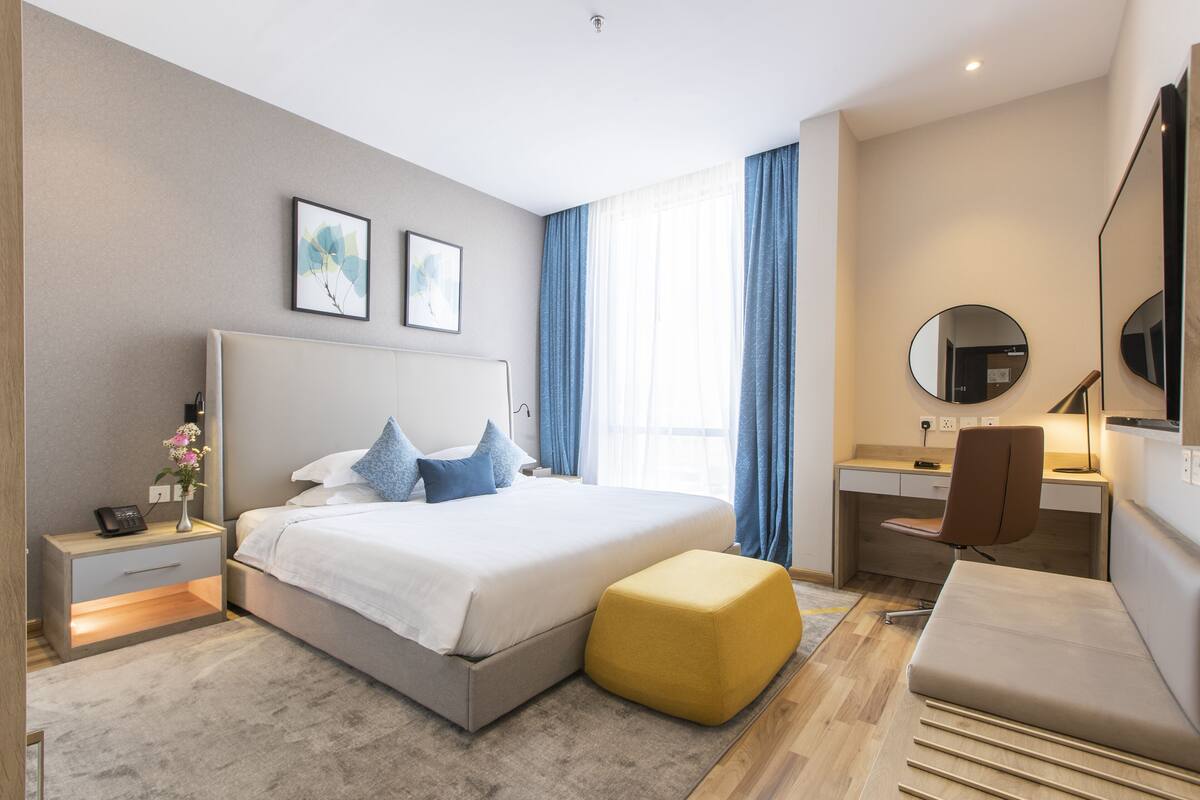A modern hotel room with a bed, bedside table, desk, chair, and a large mirror.