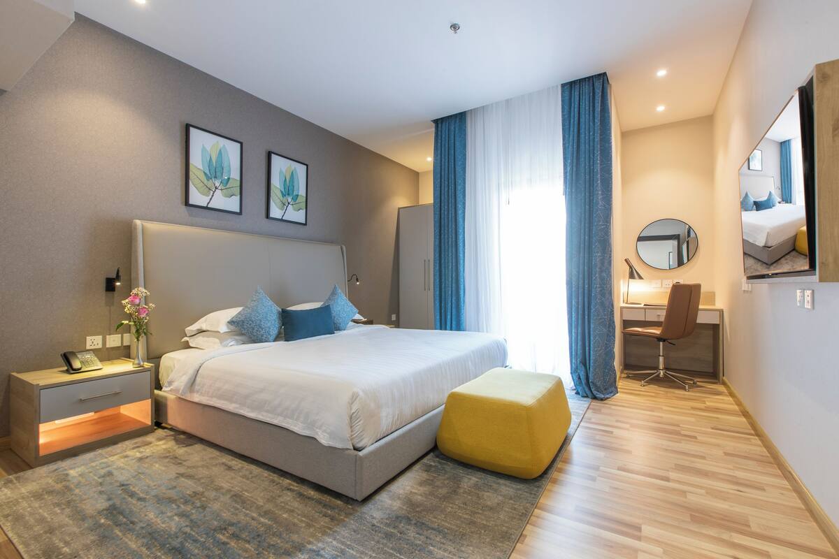 A modern hotel room with a bed, bedside table, desk, chair, and a large mirror.