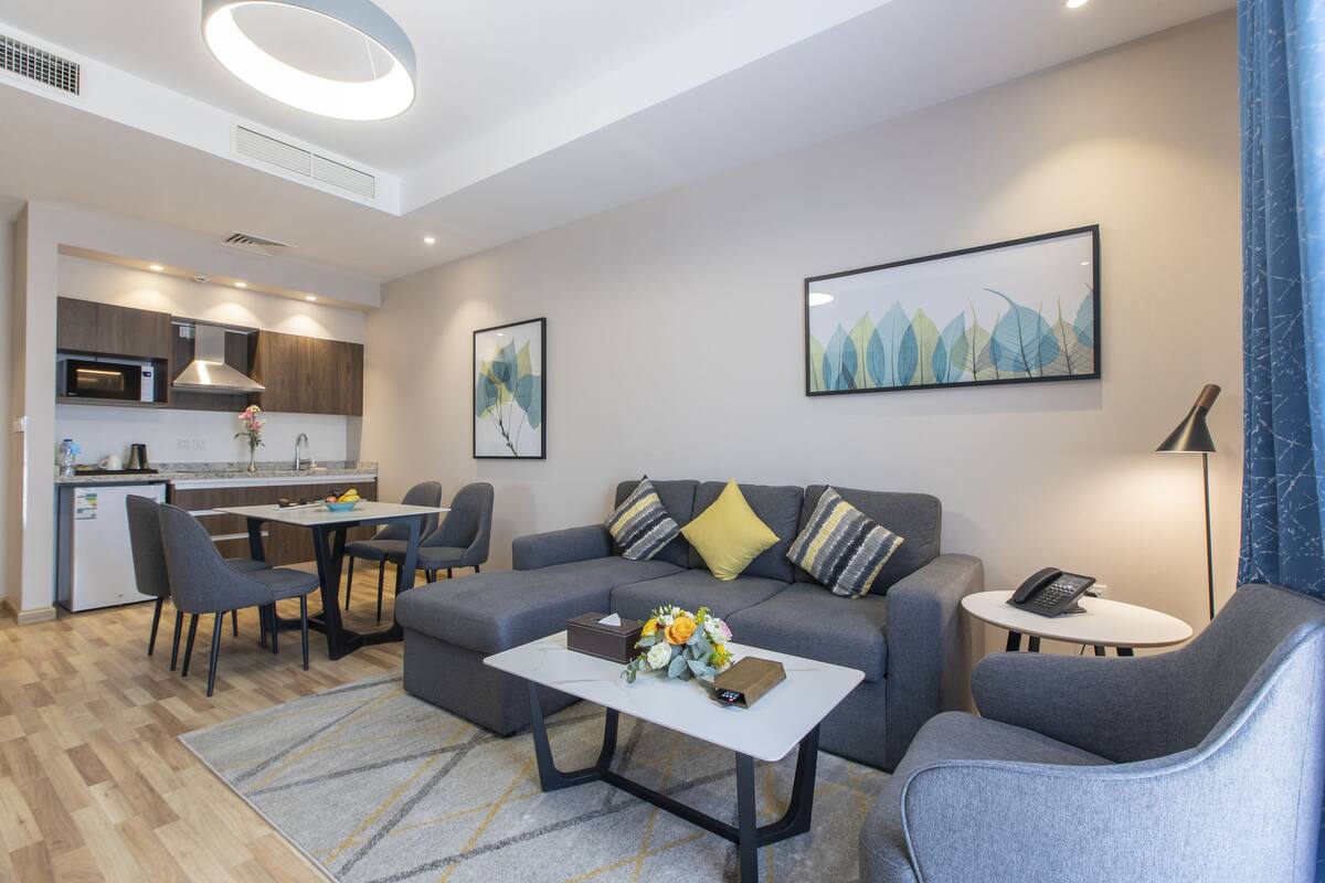 A modern living room with a grey sofa, a coffee table with flowers, a dining area with chairs, and a kitchen in the background.
