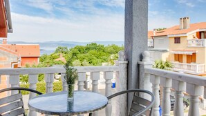 Ein Balkon mit rundem Tisch und Stühlen, einem Blumentopf sowie Blick auf das Meer und grüne Vegetation.