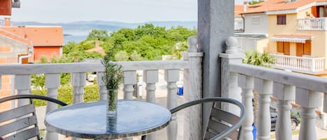 A balcony with a round table and chairs, a potted plant, and a view of the sea and greenery.