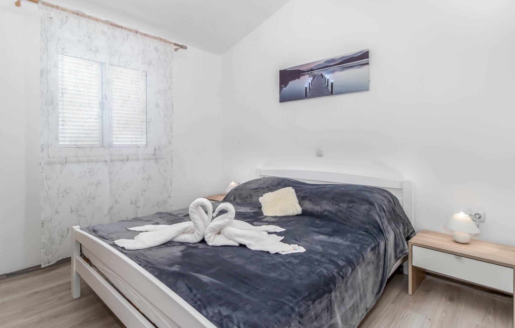 A bedroom with a bed, a nightstand, a lamp, a window with blinds, and a painting on the wall.