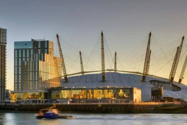 A modern stadium with a unique roof structure and multiple cranes, located near a body of water with a cityscape in the background.